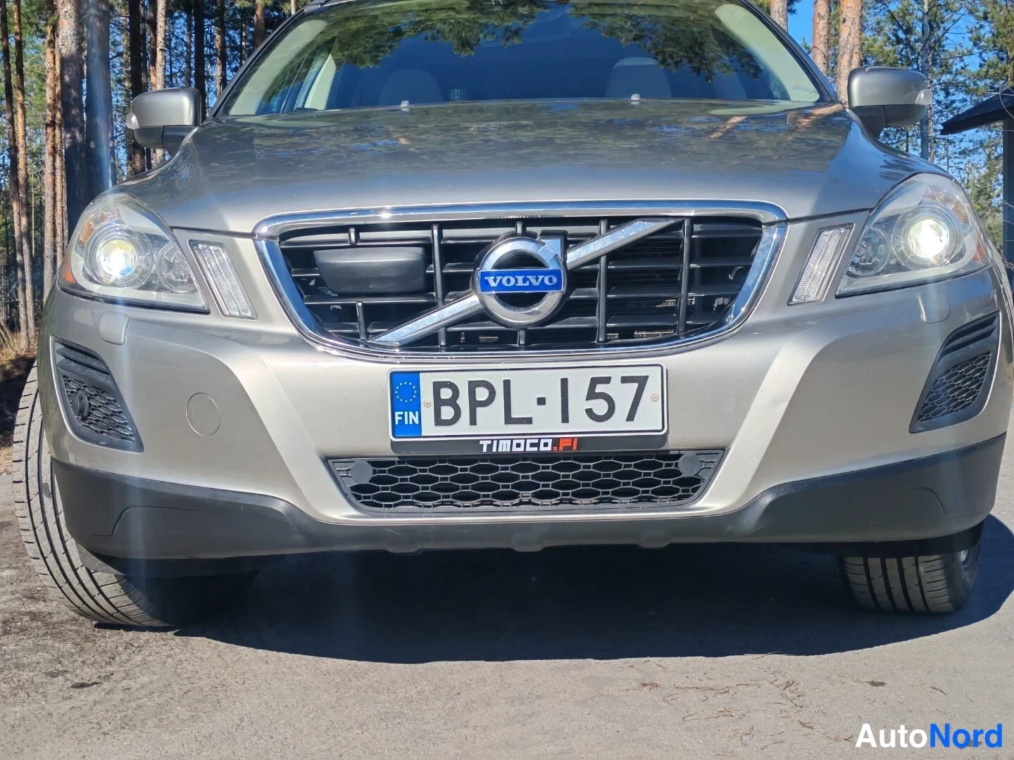 Edestä: Volvo XC60:n etupään näkymä: harmaa konepelti, VOLVO-merkki jäähdytinsuojassa, molemmat etuvalot, rekisterikilpi ja TIMOCO.FI-tunniste etupuskurissa.