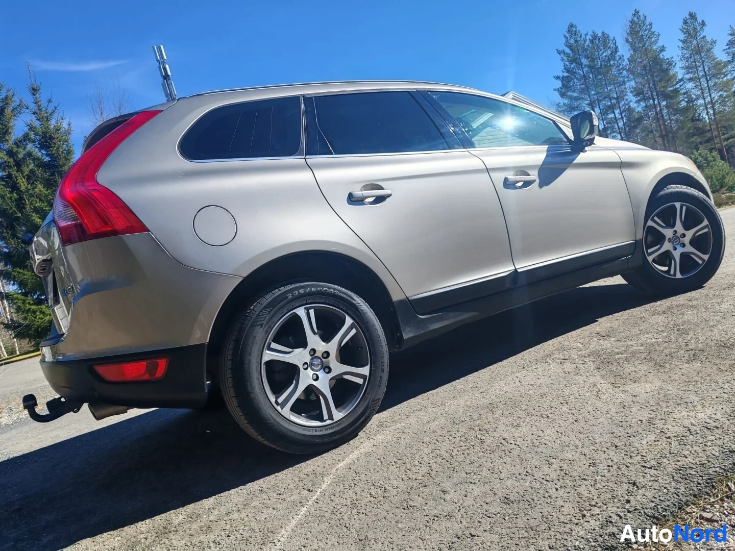 Takaviisto: Volvo XC60:n takaosakulmakuva: beigesävyinen koritakasivu ja sivu, takavalot, perävasarat ja hinauskoukku näkyvät hiekkaisella pihalla.