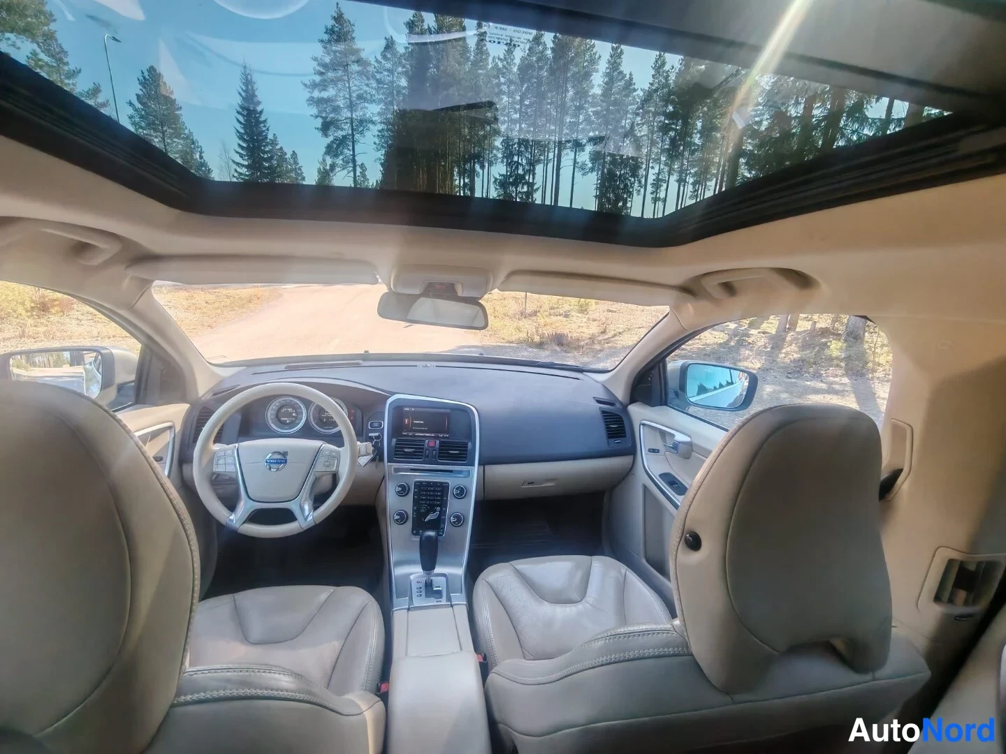 Etuviisto, nokka vasemmalle: Näkymä takaaistuimilta kohti kojelautaa: panoraamakatto, Volvo-merkkinen ohjauspyörä, näyttö keskikonsolissa ja beige nahkaistuimet.