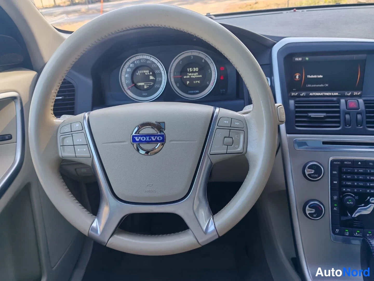 Muu: Ohjaamon näkymä: beige Volvo-ohjauspyörä, mittaristo ja keskiössä multimediajärjestelmä.
