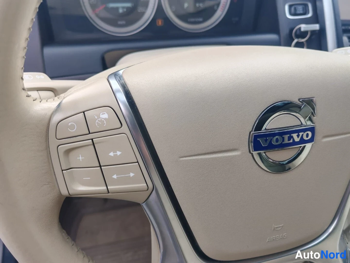 Muu: Volvo-ohjauspyörän vasen kytkinryhmä lähikuvassa, tummanpunainen logo ja kojelaudan mittarit taustalla.