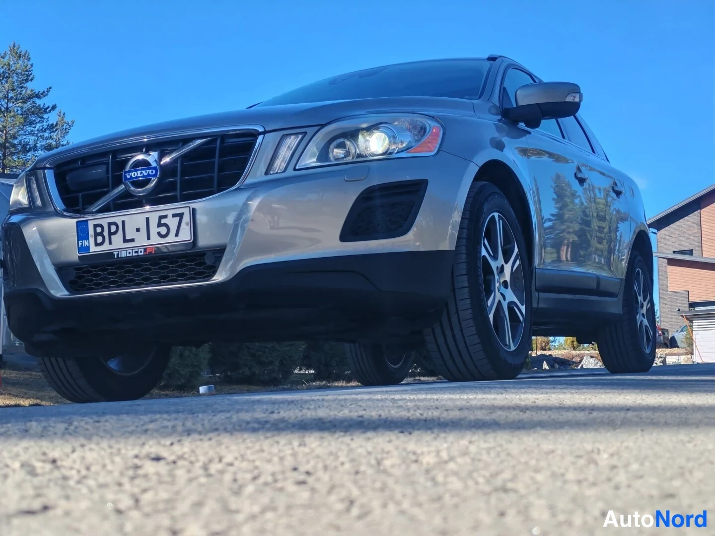 Etuviisto, nokka vasemmalle: Matalasta kuvakulmasta otettu kuva hopeanvärisen Volvo XC60:n etupäästä vasemmalta: etusäleikkö, kylki ja alumiinivanteet näkyvissä, rekisterikilpi lue