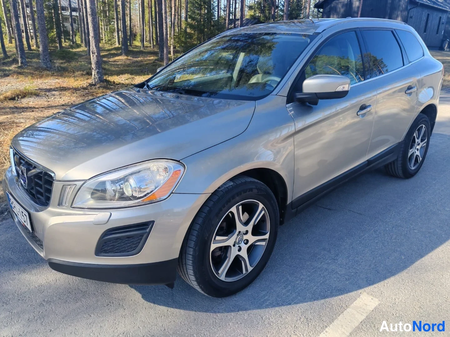 Etuviisto, nokka vasemmalle: Kulmakuva hopeanvärisen Volvo XC60:n etupäästä vasemmalta, koko kylki näkyvissä: kattokaiteet, mustat ikkunakehykset ja alumiinivanteet, rekisterikilpi