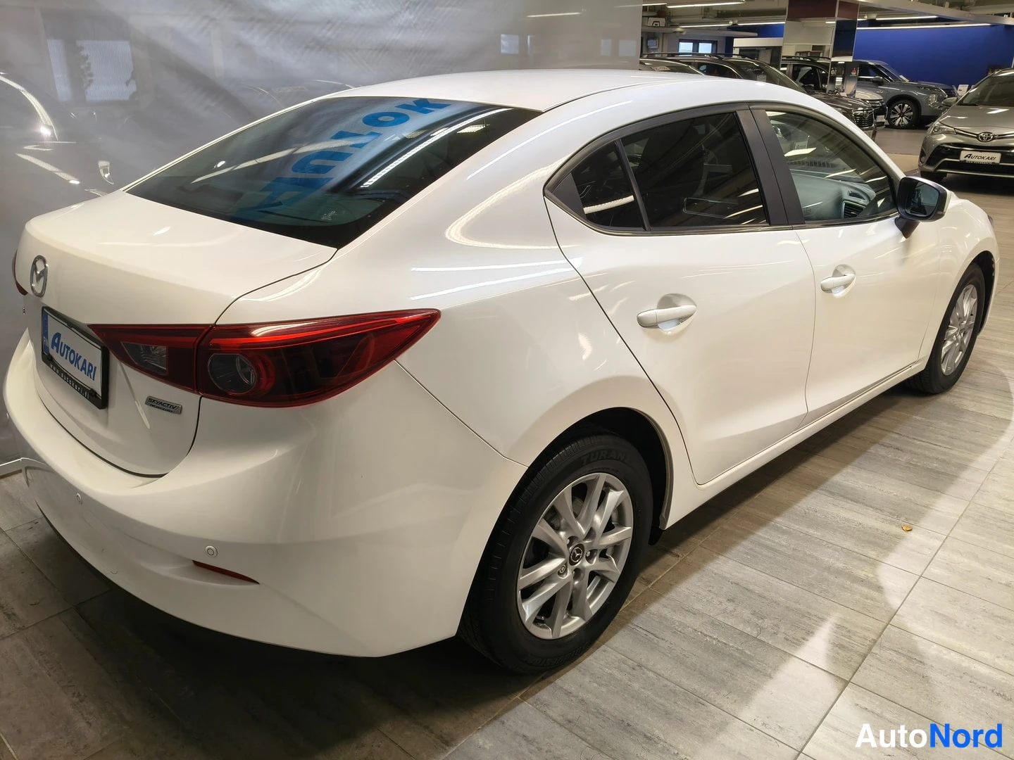 Takaviisto: Valkoinen Mazda3-sedan takaviistosta autosalissa; koko korimalli näkyy, takavalot ja ovien kahvat selkeästi näkyvissä.
