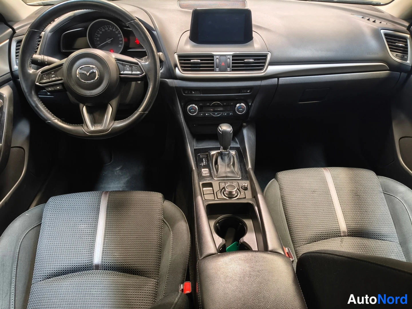 Muu: Mazda3:n ohjaamo: musta nahkapinnetty ohjauspyörä, mittaristo, keskinäytin, automaattivaihteiston valitsin ja kangaskojobussit.