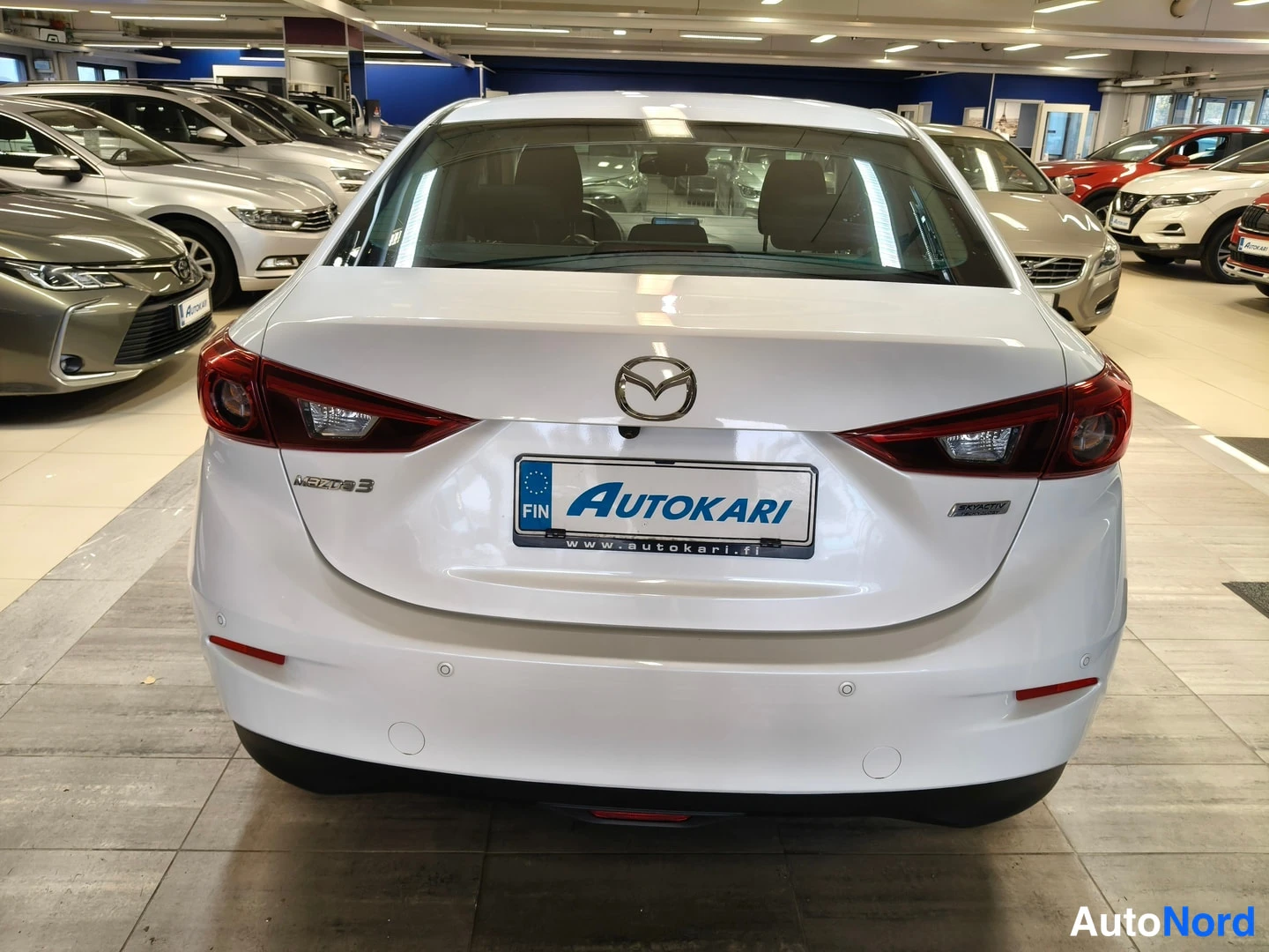 Takaa: Mazda3-sedanin perä suoraan takaa; valkoinen maali, punaiset takavalot, Mazda-merkki ja Autokari-tunnuksella varustettu rekisterikilpi.