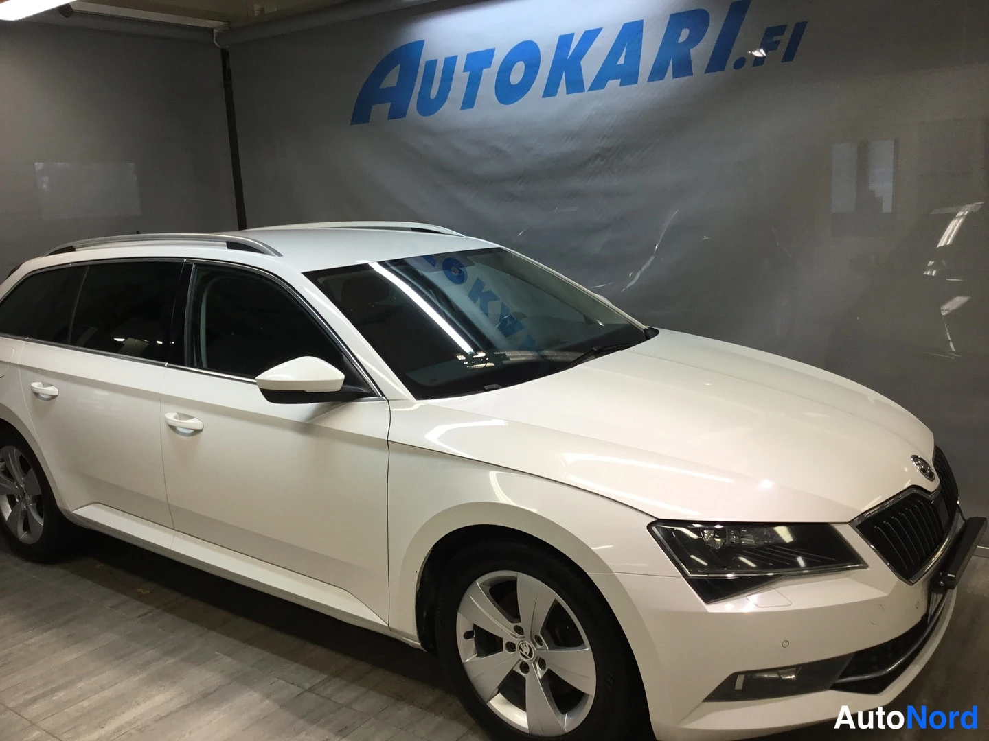 Etuviisto, nokka oikealle: White estate car photographed front three-quarter from the nose-right in a showroom, roof rails and alloy wheels visible.