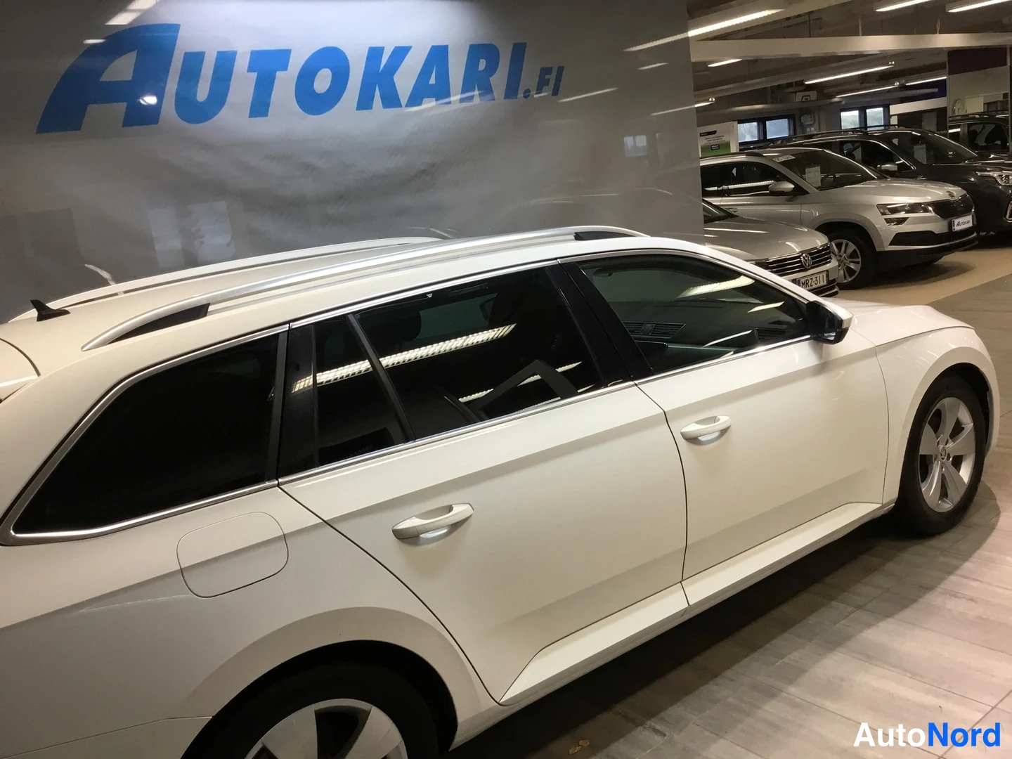 Sivulta: White estate car photographed from the side in a showroom, showing doors, tinted windows and alloy wheels.