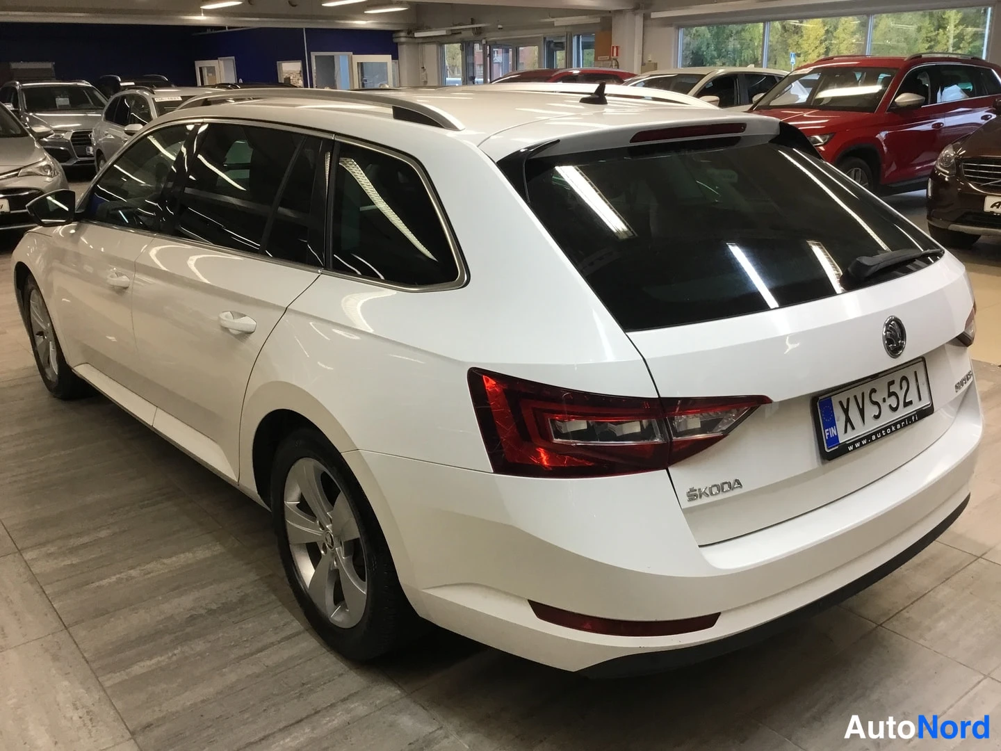 Takaviisto: White estate car photographed rear three-quarter from the tail-left indoors, tail lights, boot lid and license plate visible.