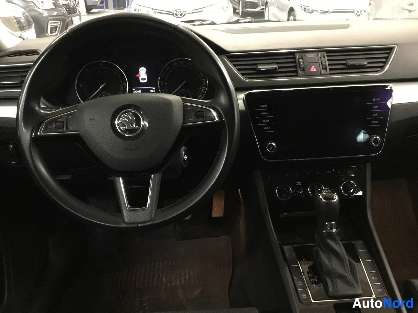 Edestä: Ajonäkymä Skoda Superbin ohjaamosta: ohjauspyörä, mittaristo, keskikonsolen infotainment-näyttö ja vaihtevalitsin.