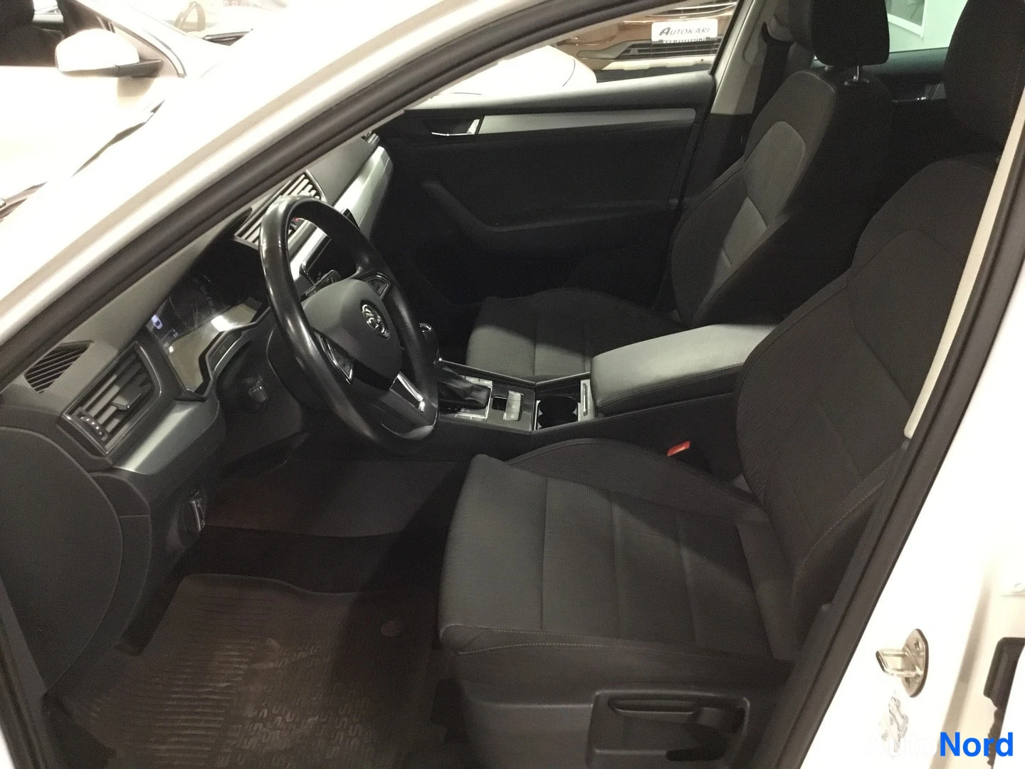 Edestä: Skoda Superbin etuistuinriviltä näkymä ohjaamoon: mustat kankaanpenkit, ohjauspyörä, keskikonsoli ja ovi auki.