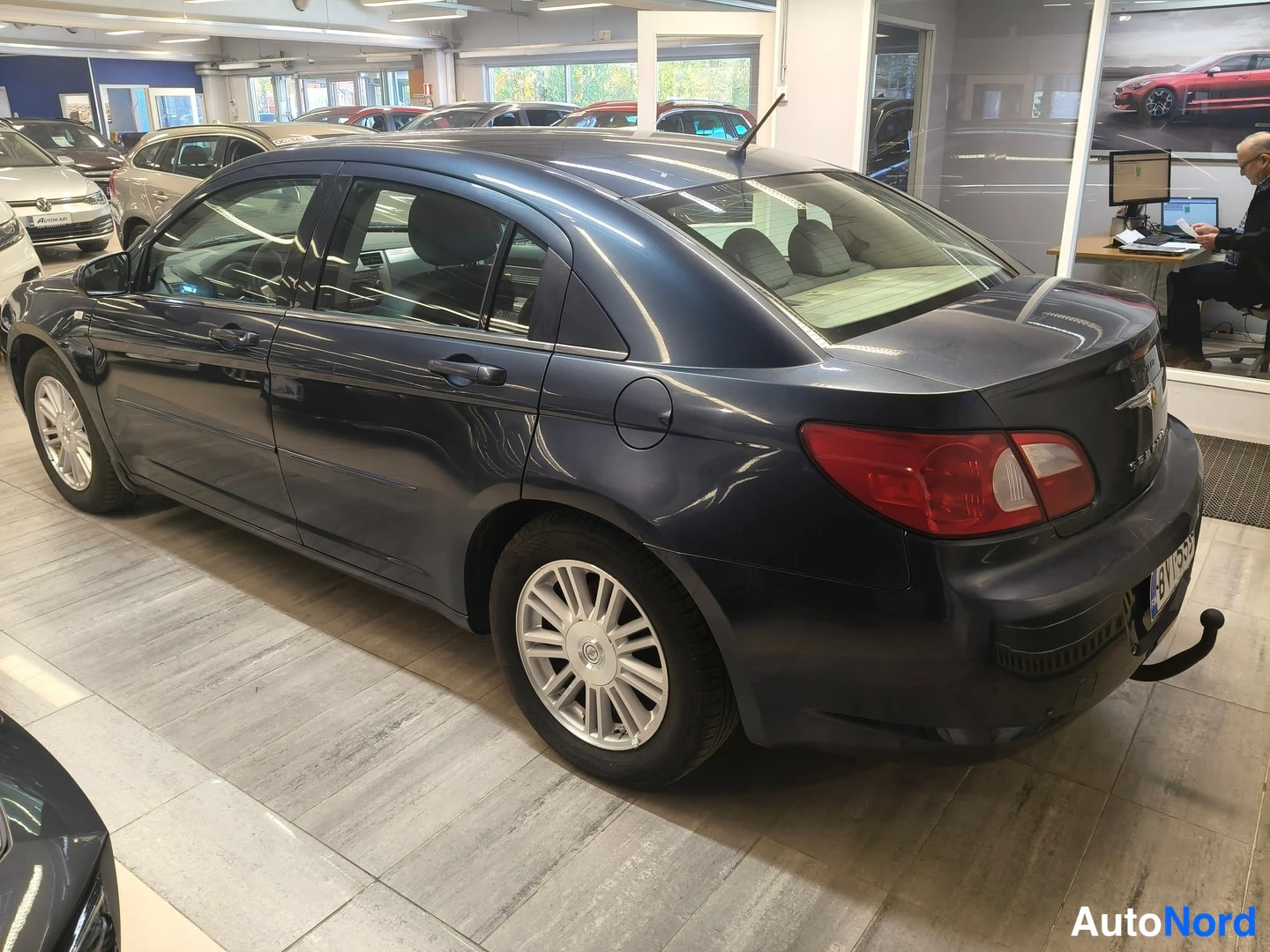 Takaviisto: Tummansininen Chrysler Sebring -henkilöauto kuvattuna takaviistosta autosalissa; takavalot, tavaratilan kansi ja kylki näkyvät, takaosassa vetokoukun kaula, rek