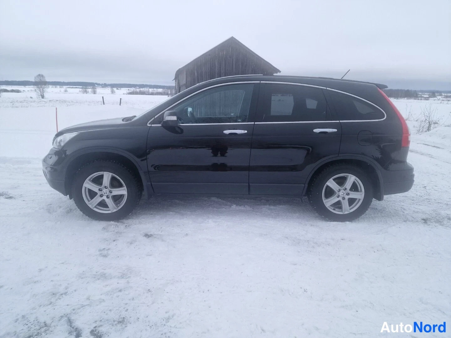 Sivulta: Mustan Honda CR-V:n sivukuva oikealta puolelta lumikentällä, taustalla hirsilato.