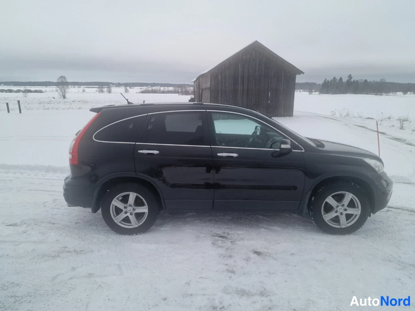 Sivulta: Musta Honda CR-V -farmarityyppinen maastokuvaus sivusta lumen peittämällä pihalla, talli taustalla.