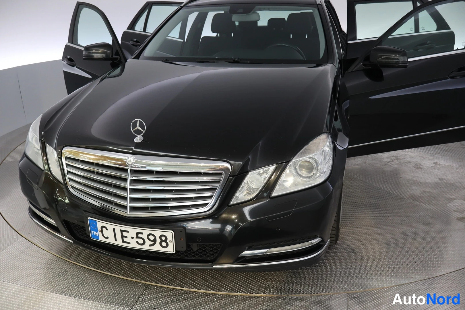 Etuviisto, nokka vasemmalle: Mustan Mercedes-Benzin keula kolmeneljänneskuvassa oikealta, keulakoriste ja rekisterikilpi näkyvät, ovien ollessa auki.