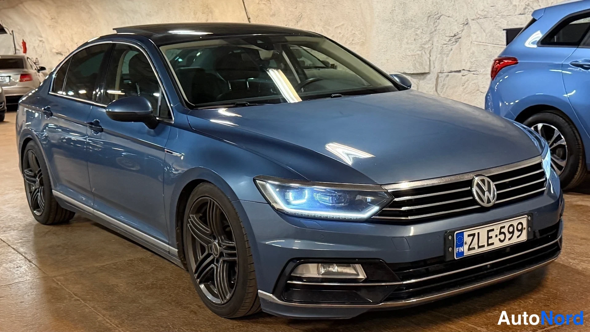 Etuviisto, nokka vasemmalle: Sininen Volkswagen Passat -sedan edessä kolmannesvasemmalla kuvakulmasta, LED-etuvalot päällä, rekisteri.