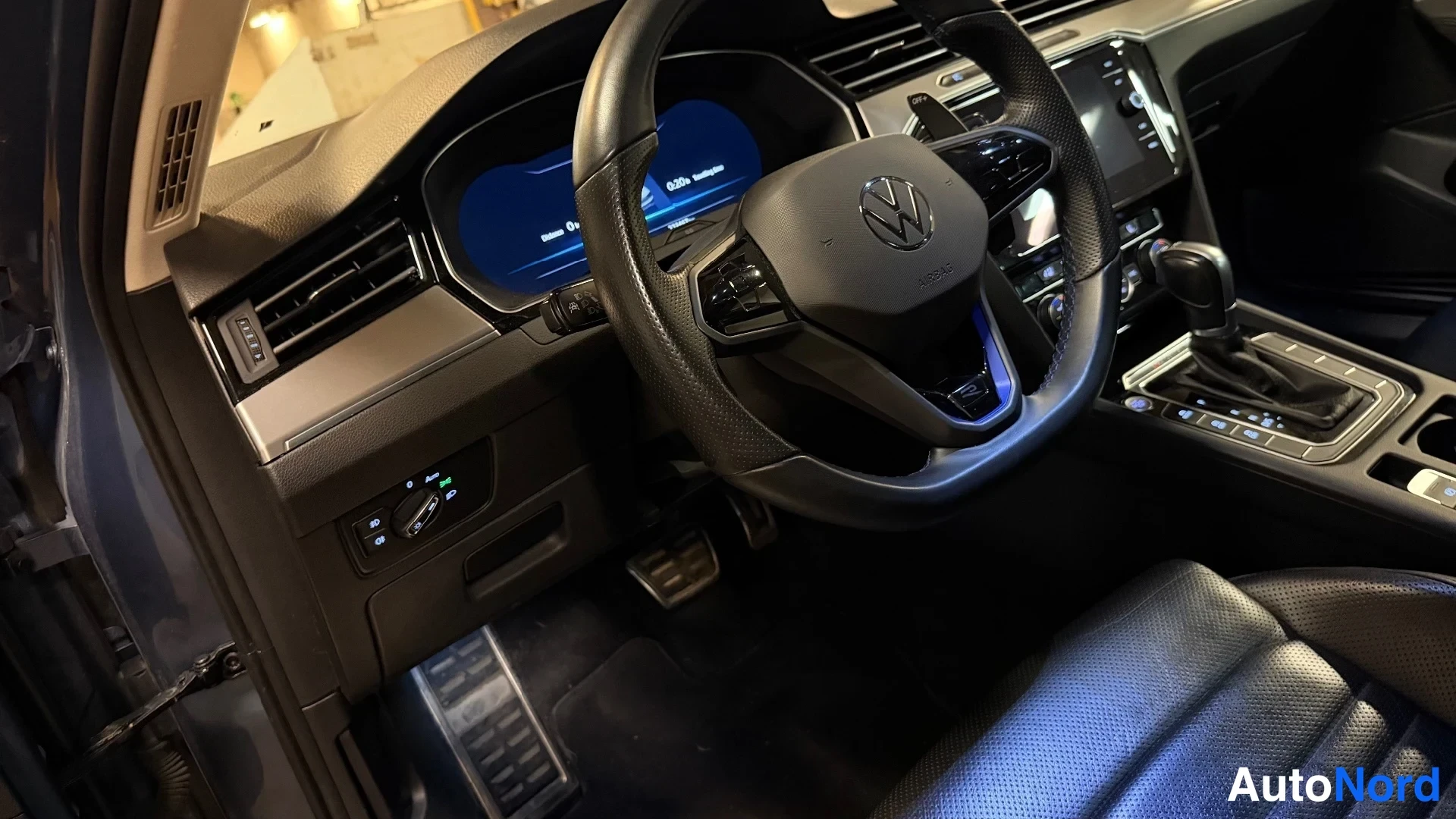 Yksityiskohta: Volkswagenin etusisusta kuljettajan puolelta: tumma nahkainen ohjauspyörä ja VW-merkki, sisäpuolella näkyy nopeusmittari, vaihteenvalitsin, polkimet ja ikkunano