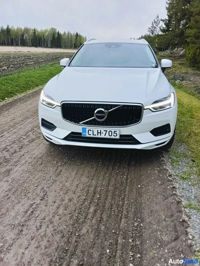Edestä: Valkoinen Volvo XC60 suoraan edestä päin, etusäleikkö ja rekisterikilpi näkyvät hiekkatiellä.