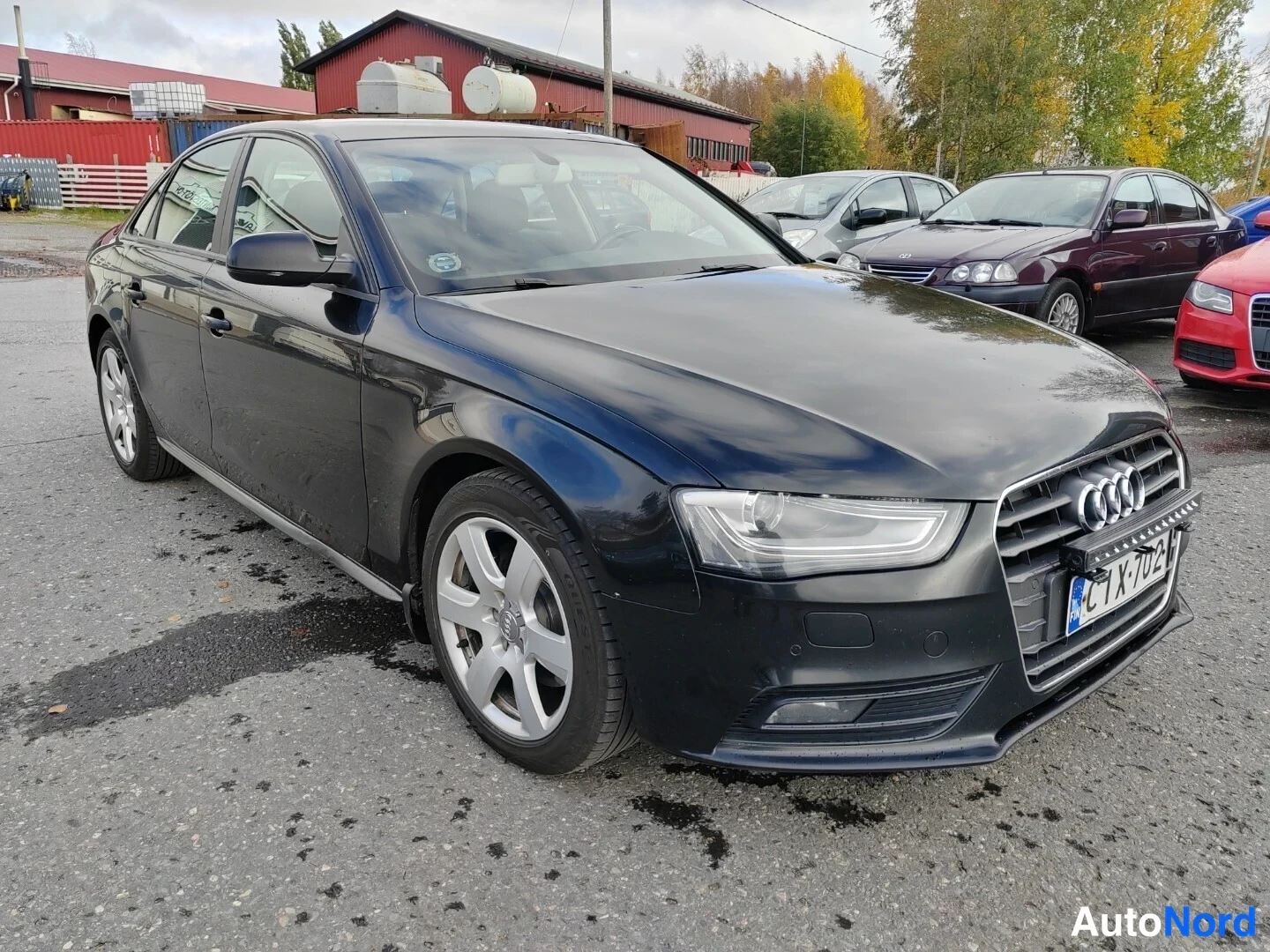 Etuviisto, nokka oikealle: Mustanharmaan sävyinen neliovinen Audi-sedan kuvattu etukulmasta oikealta; etusäleikkö ja Audi-merkki, rekisteritunnus, hopeat peltivanteet.