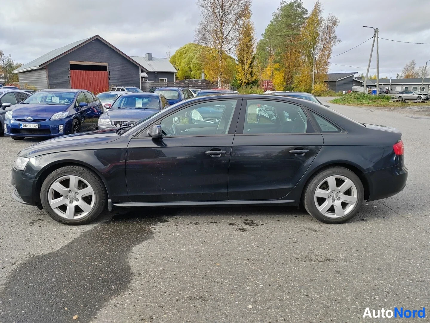 Sivulta: Musta Audi A4 neliovisinen sedani kuva autosta sivulta. Auto seisoo asfaltilla ja sen vieressä on muitakin autoja taustalla.