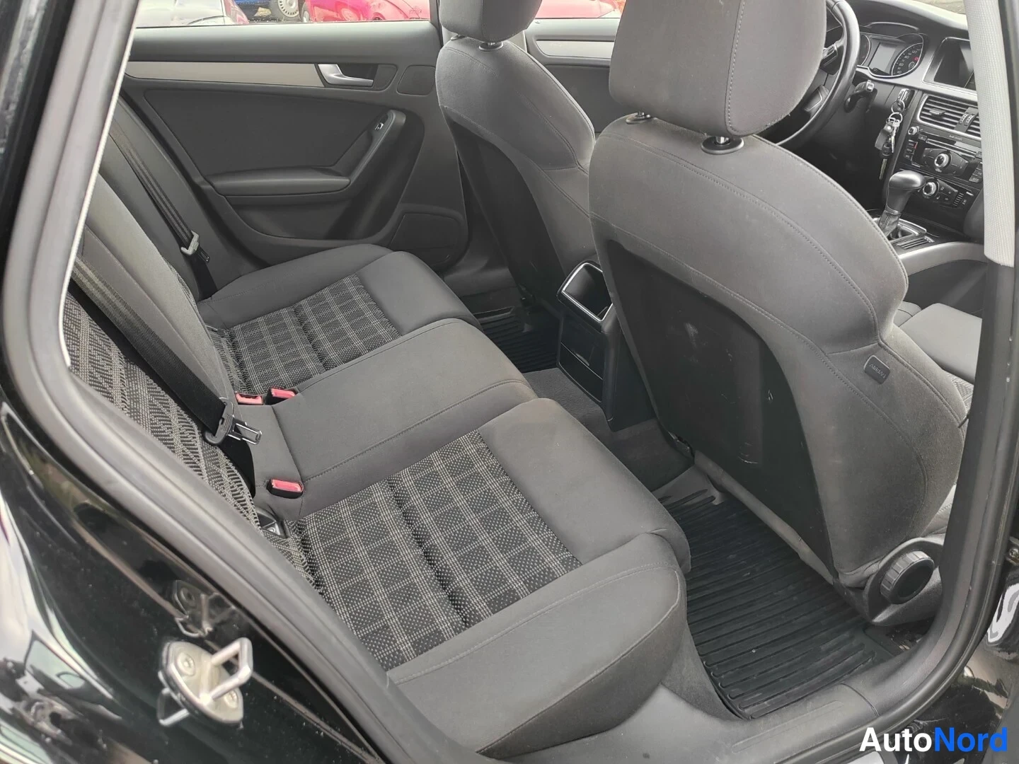Takaistuimet: View of the car's interior rear seats with a front seat back in view. The back seat is black fabric with grey plaid inserts. The front seats are solid black clo