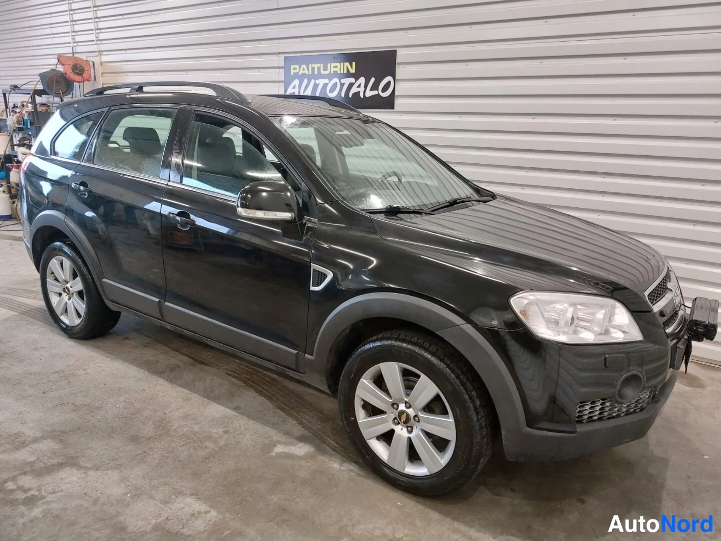 Etuviisto, nokka vasemmalle: Musta Chevrolet Captiva -katuauton etu- ja vasen sivunäkymä sisätilassa. Auton pinnalla heijastuu valoja. Takavalot ja ajovalot näkyvät selkeästi.
