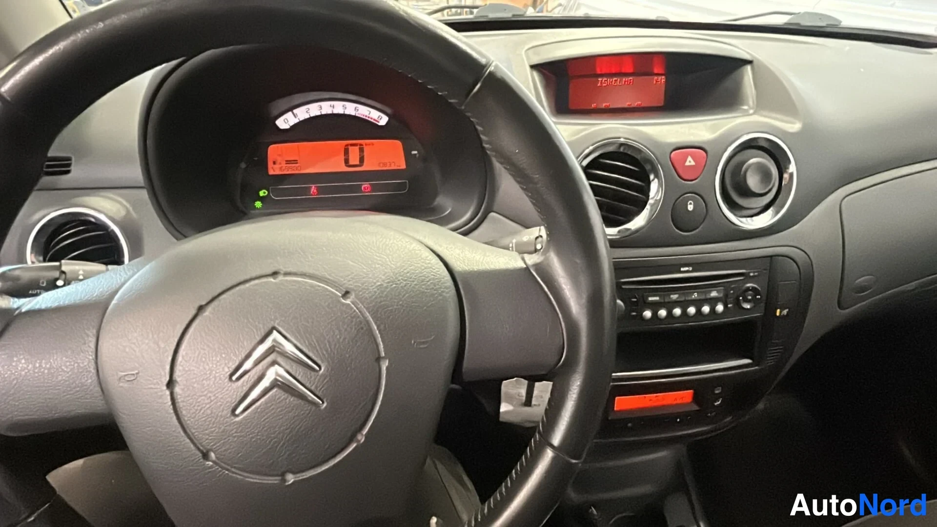 Muu: Citroenin ratin ja kojelaudan lähikuva; mittaristo näyttää 0 ja keskikonsolissa radio ja ilmastointia tai lämpöä 'LISÄLÄMMA' -tekstillä.