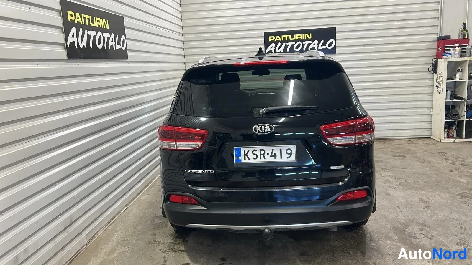 Takaa: Kia Sorenton suora takanäkymä sisätilassa: mustat kiiltävät koripanelit, takavalot, KIA-merkki ja Sorento-teksti takaluukussa, rekisterikilpi, kattokaiteet sekä