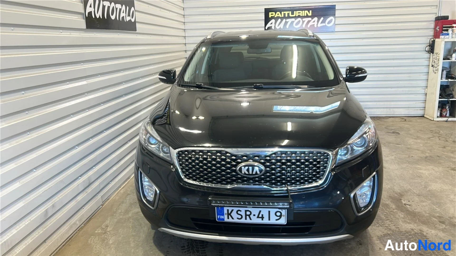 Edestä: Kia Sorento -maasturin suora etunäkymä sisätiloissa; tumma kori, KIA-merkitty etusäleikkö ja rekisterikilpi näkyvät.