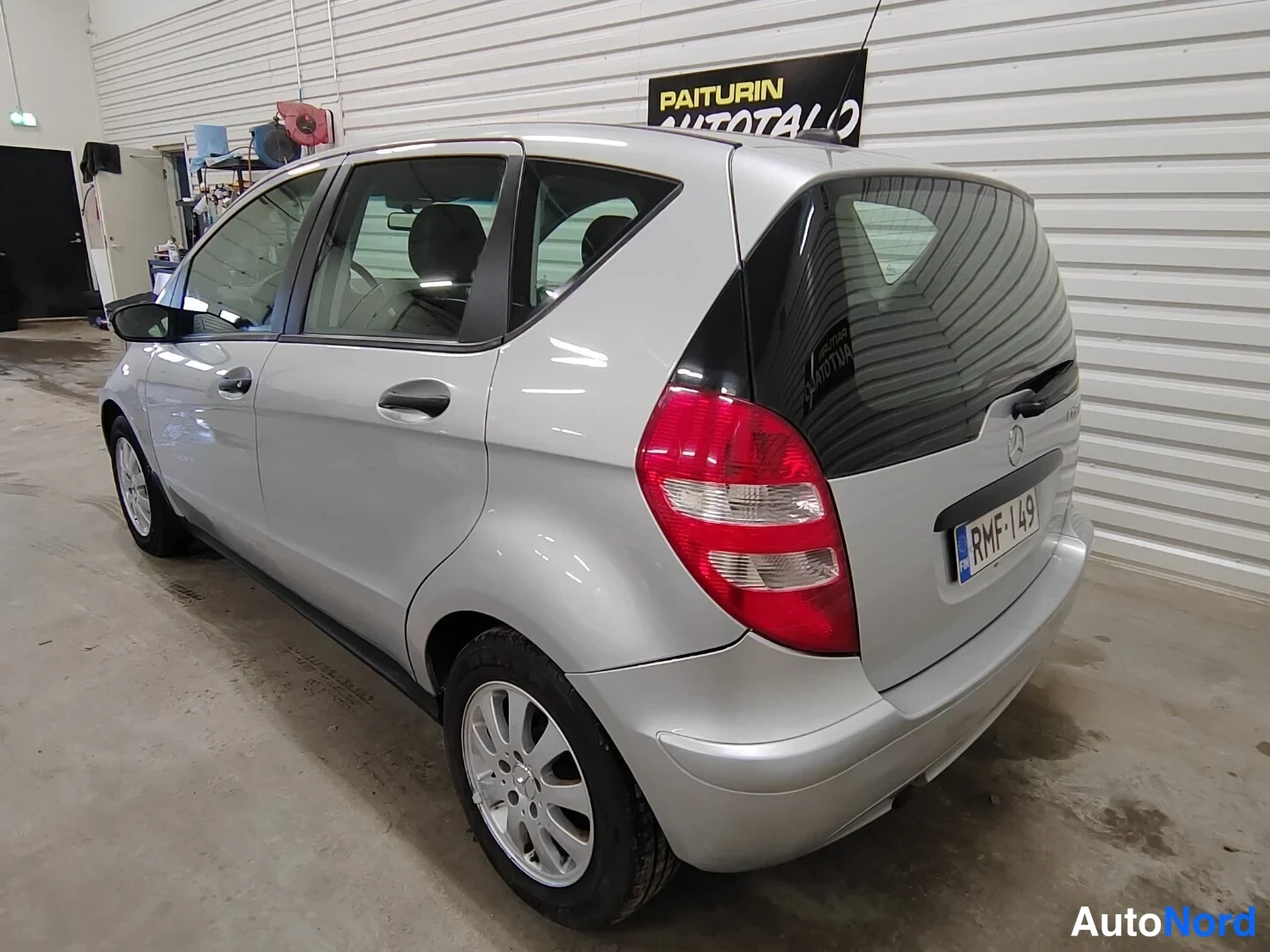 Takaviisto: Silver five-door hatchback shown from a rear three-quarter angle inside a garage with corrugated white walls.