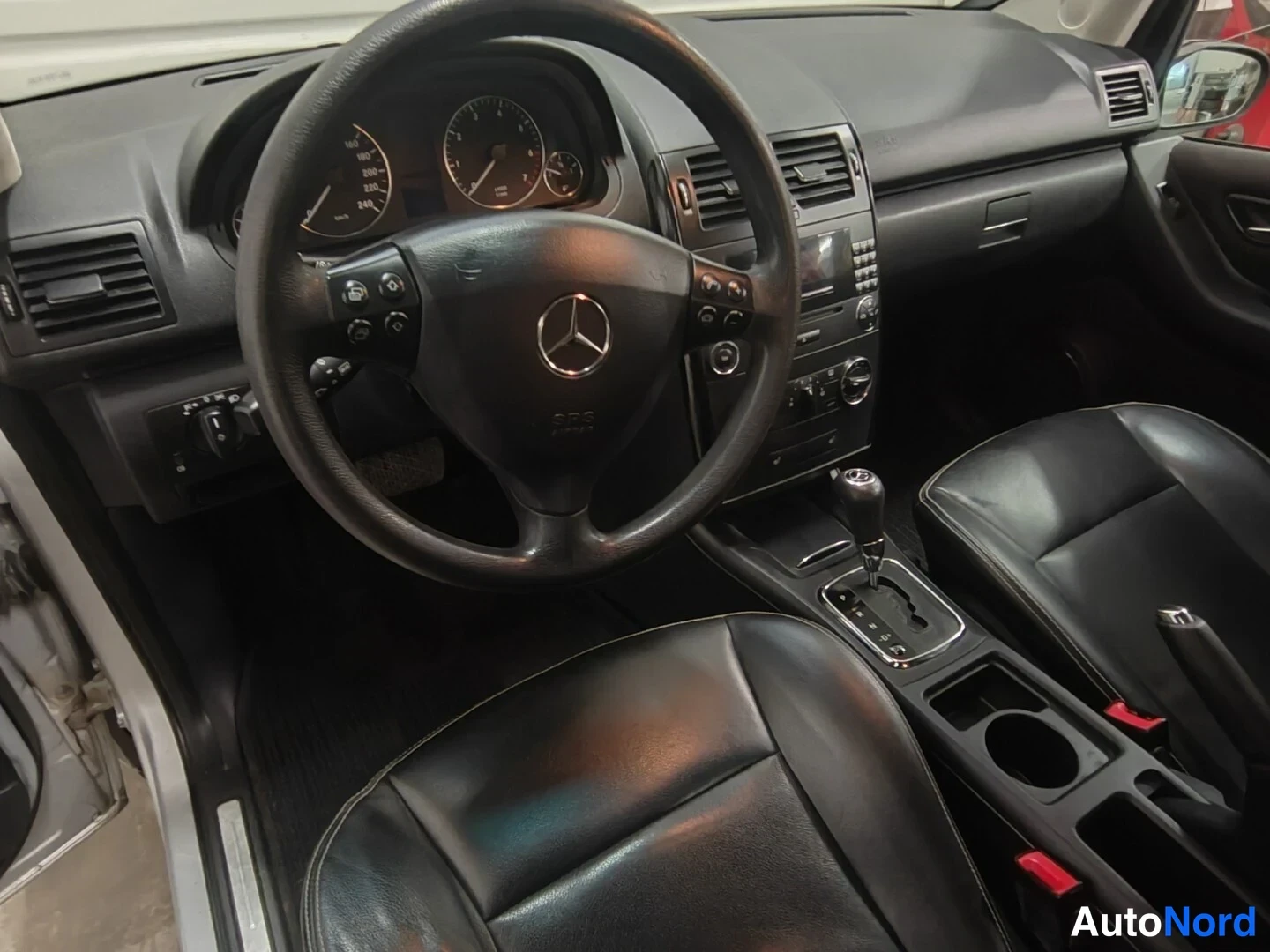 Muu: Ohjaamo vasemmalta ohjattavasta Mercedes-Benzistä: Mercedes-merkitty ohjauspyörä, analogiset mittarit ja automaattivaihteen valitsin keskikonsolissa, mustat nah