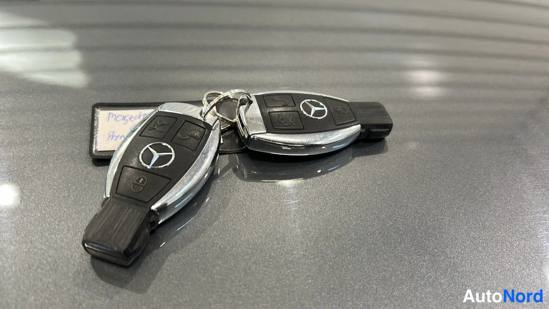 Yksityiskohta: Kaksi Mercedes-avainnippua Mercedes-tähtimerkeillä metallinhohtoisen harmaan pinnan päällä; takana osittain näkyvä käsin kirjoitettu lappu.