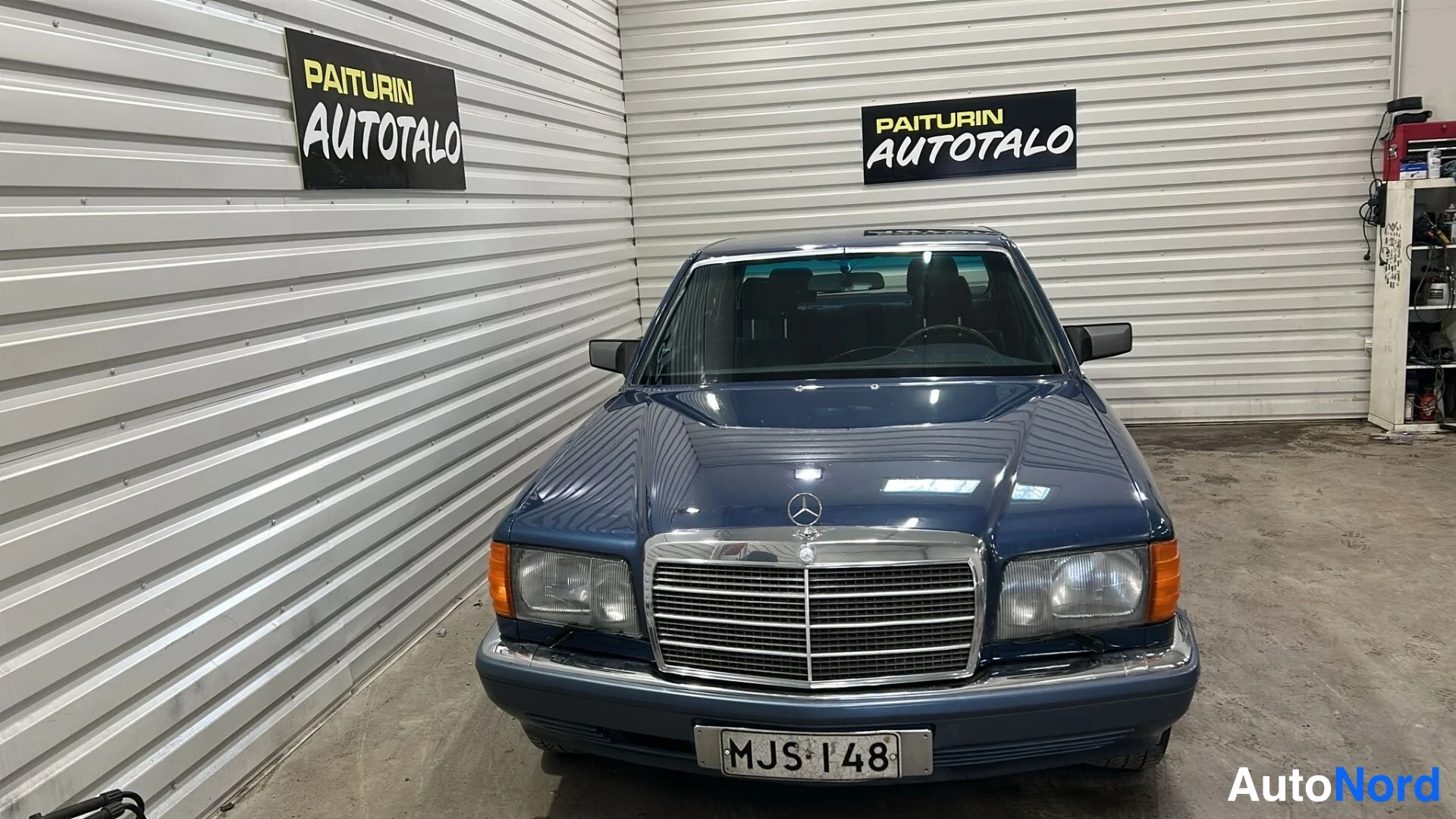 Edestä: Sininen Mercedes-Benz sedan suoraan edestä, konepellin merkki, krominen etusäleikkö ja pyöreät ajovalot näkyvät.