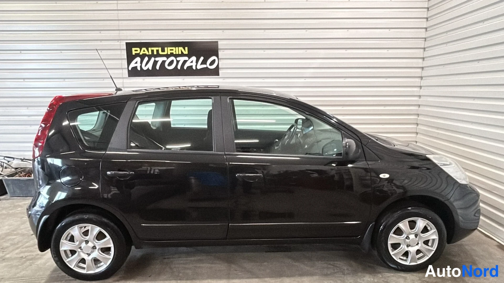 Etuviisto, nokka oikealle: Musta Nissan Note -henkilöauto sivuprofiilista oikealta hallissa.