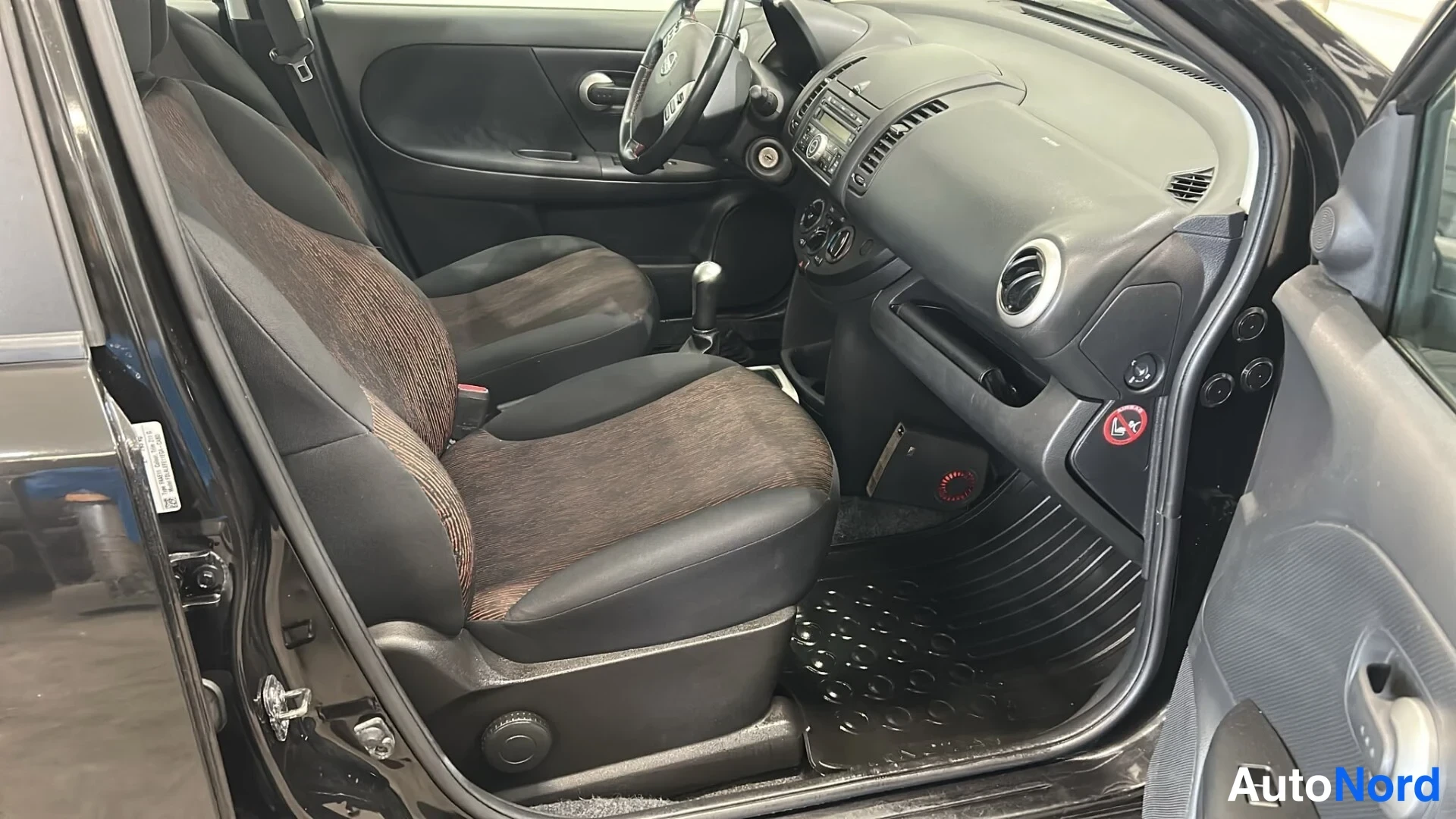 Muu: Avoimesta kuljettajan ovesta kuvattu Nissan Noten sisätila: etusomit, kojelauta, voimansiirtokeppi ja mustat tekstiilimatot.