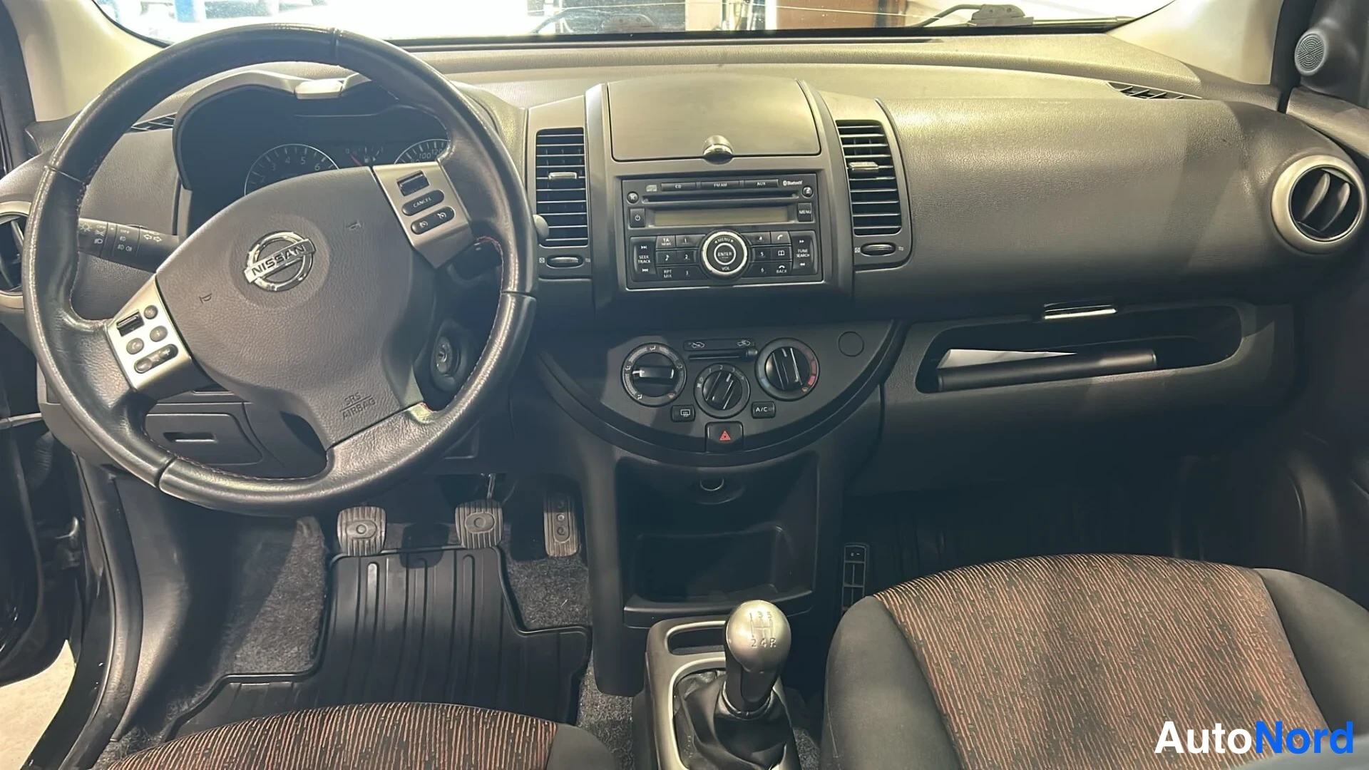 Edestä: Ohjaamo näkyy suoraan etualalta: Nissan-ohjauspyörä, keskellä radio ja ilmastointisäätimet, manuaalivaihteen keksi, tummat matot ja ruskeakuvioinen istuin.
