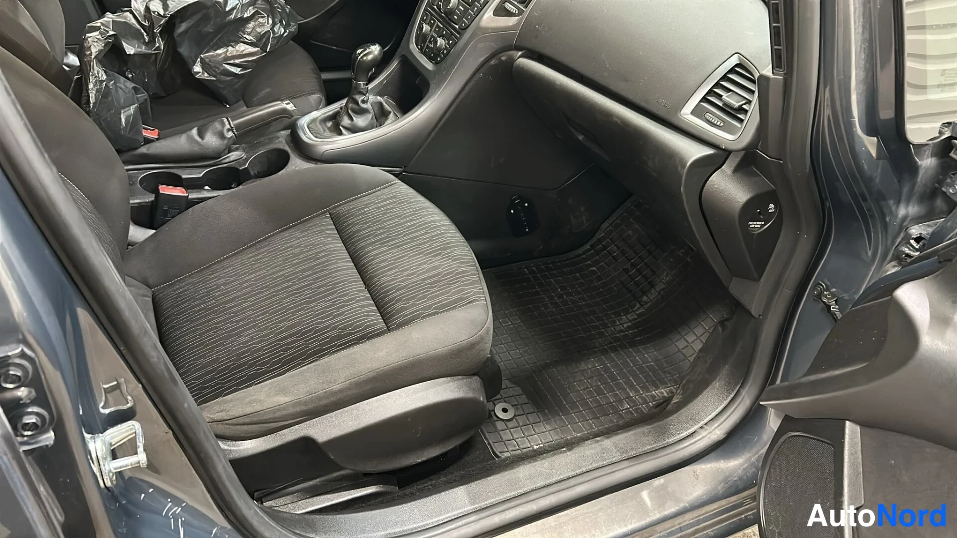 Muu: Sisäkuva etuistuimesta ja mittaristosta: musta kangasistuin, manuaalivaihteisto ja lattiakaavaimet näkyvät avoimesta ovenraosta.