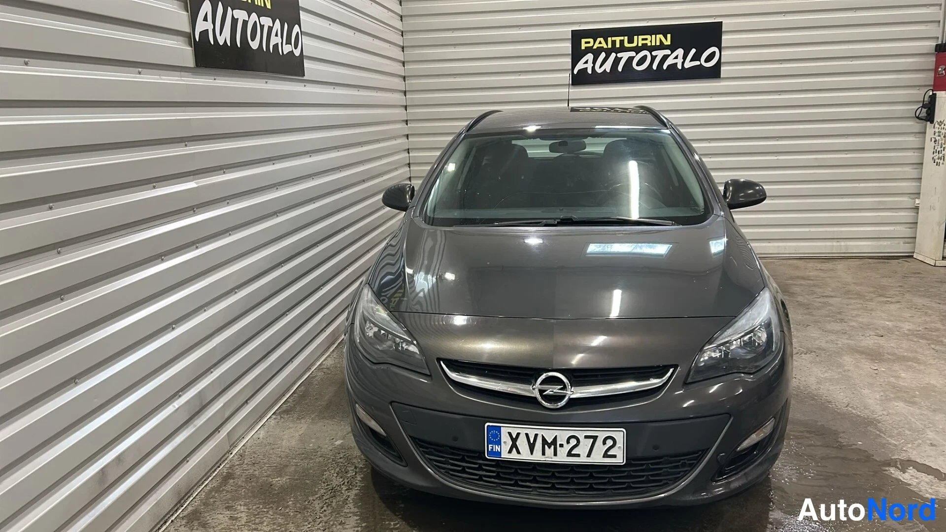 Edestä: Tummanharmaan farmariauton keulakuva suoraan edestä sisätiloissa; etuvatopisset, Opel-merkki ja rekisterikilpi näkyvät.