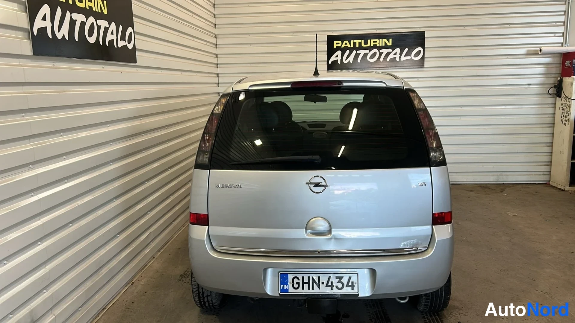 Takaa: Auton takaosa opastettuna: hopeanvärinen farmarimallinen henkilöauto, rekisteritunnus, Opel-merkki ja Meriva-teksti takaluukussa.
