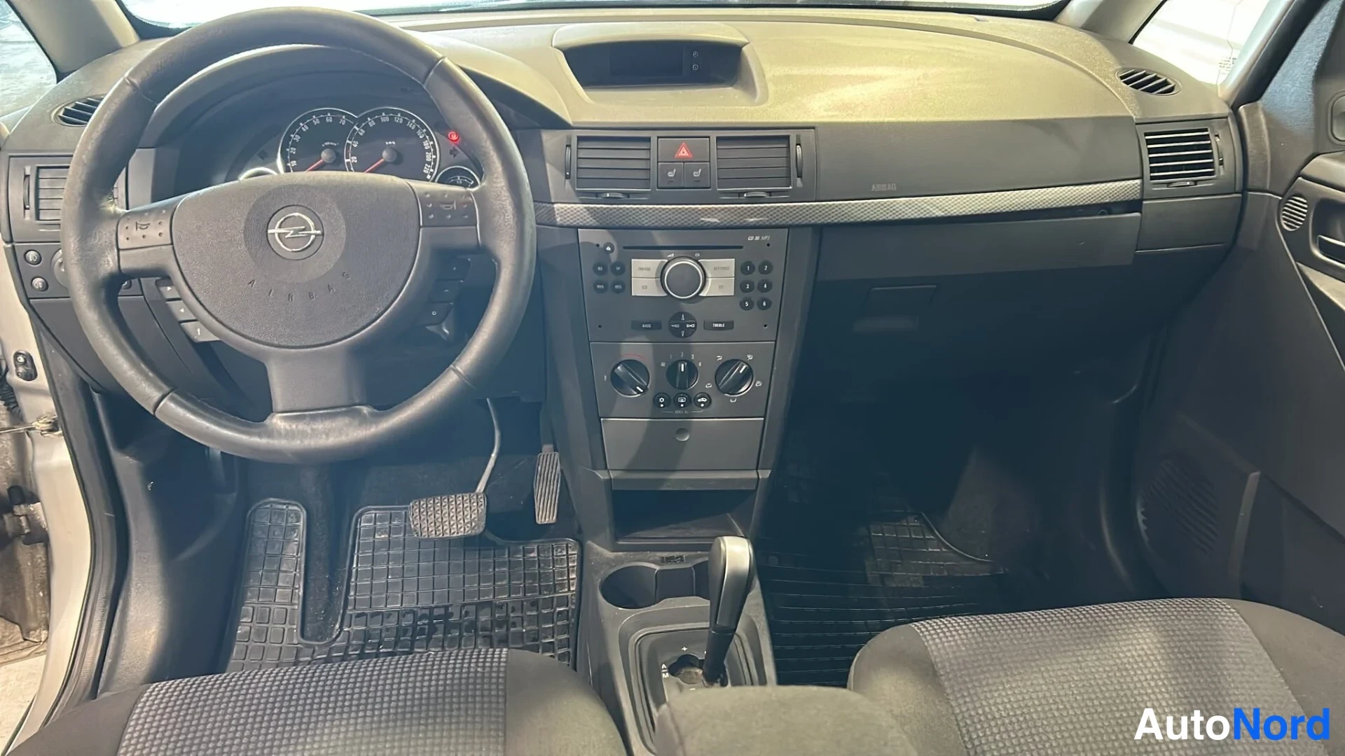 Edestä: Ohjaamo näkyy etupuolelta: Opel-merkkinen ohjauspyörä, mittaristo, keskiöpaneeli radio- ja kierrätys控件illä ja automaattivaihteen valitsin, mustat matot lattiall