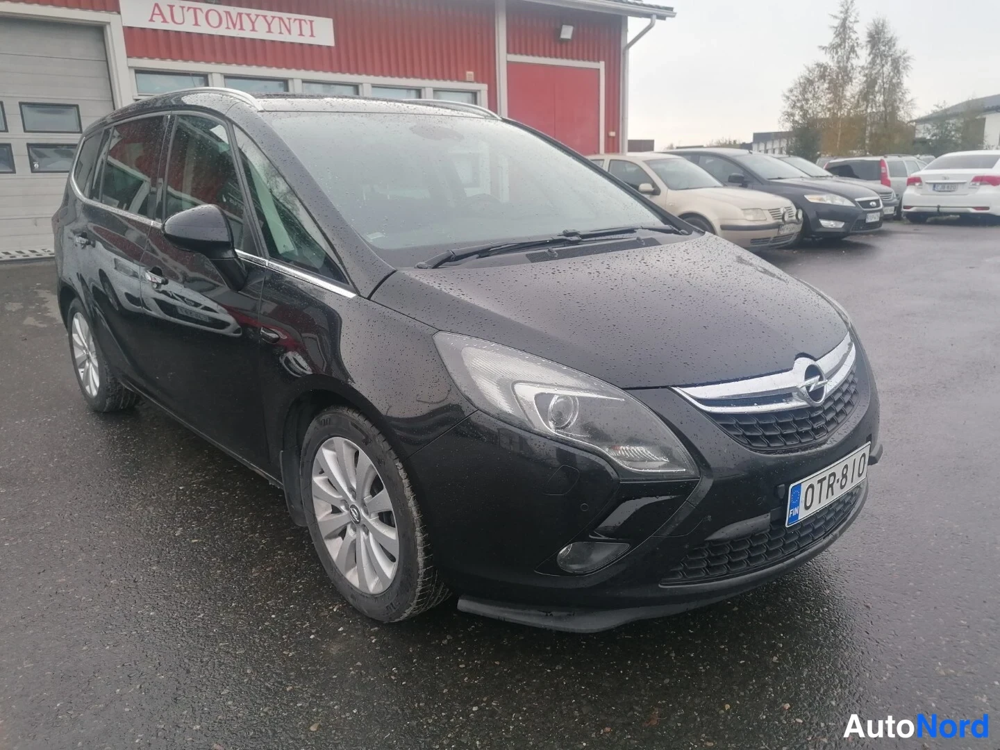 Etuviisto, nokka oikealle: Musta Opel Zafira Tourer vinosti edestä oikealta, rekisterikilpi, kattokiskot ja hopeiset monipeltovanteet, sadepisaroin peitetty kori, taustalla punaine