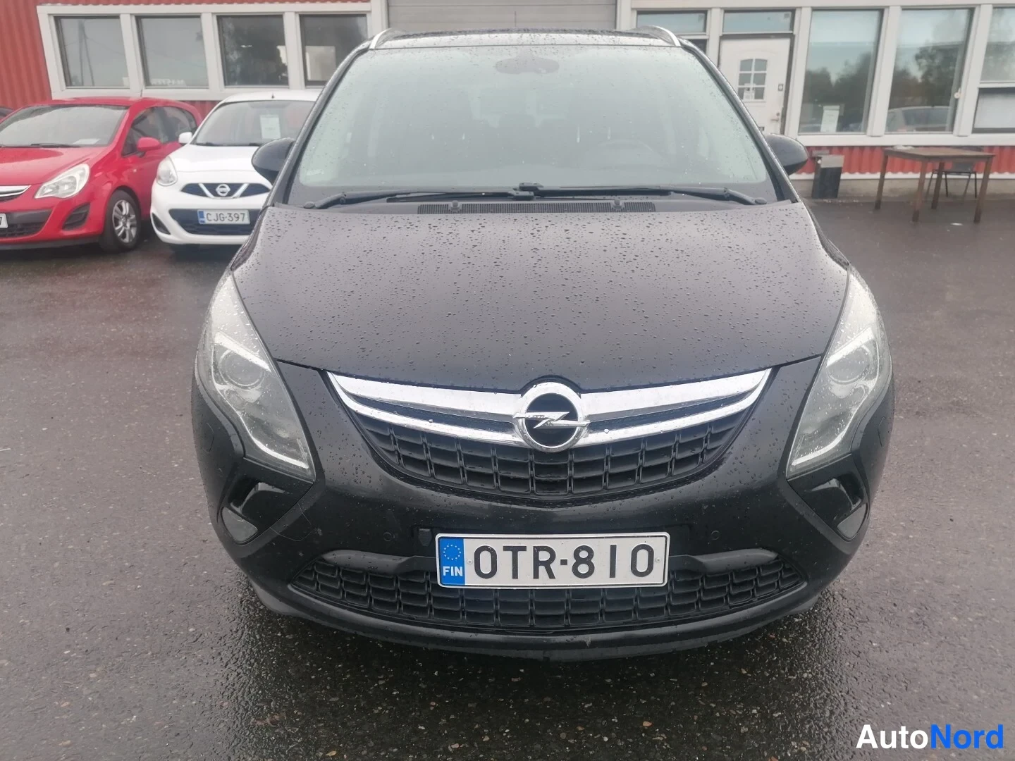 Edestä: Mustan Opelin suora etänäkymä, kromilistainen etusäleikkö ja Opel-logo, rekisterikilpi FIN, märkä asfaltti, taustalla punainen ja valkoinen henkilöauto.