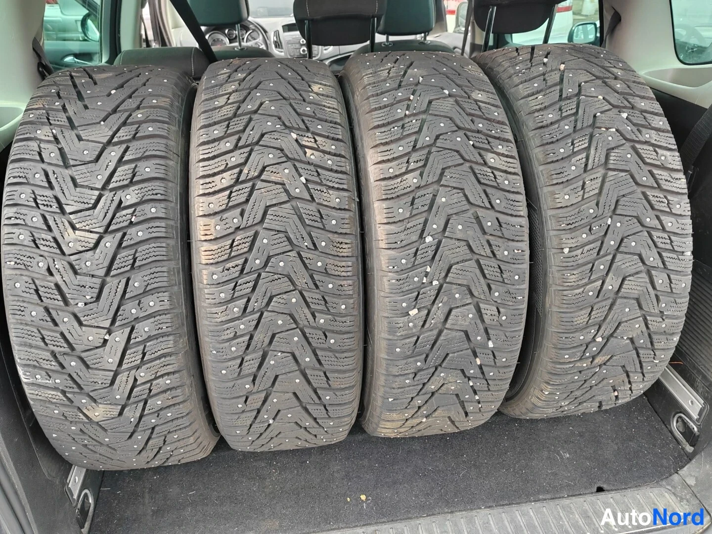Muu: Neljä nastarengasta vierivällä kuvioilla seisoo rivissä autossa matolla lattialla.
