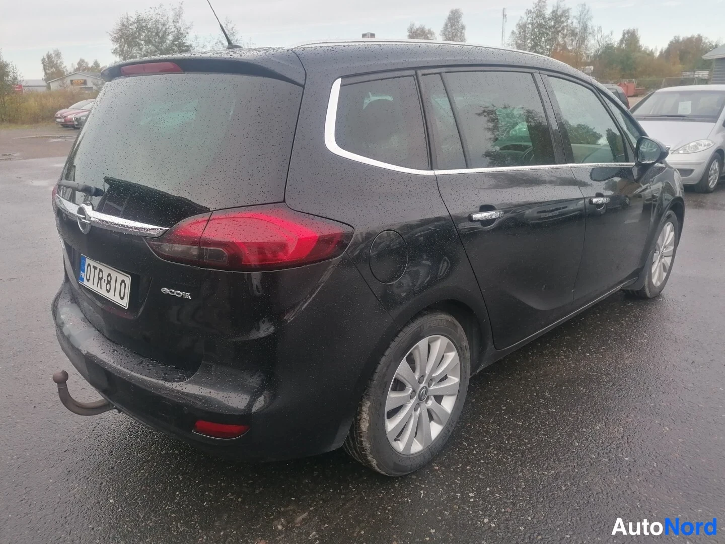 Takaviisto: Mustan Opel Zafiran takakolmannes vasemmalta, punaiset takavalot, ecoFLEX-merkki, rekisterikilpi ja vetokoukun pallokytkin, taustalla hopeinen pakettiauto.