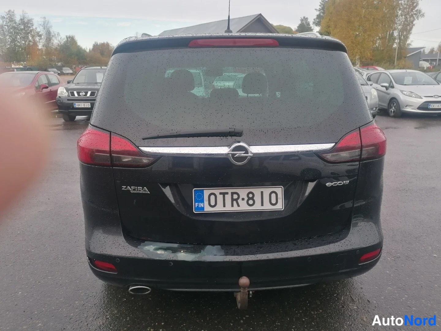 Takaa: Black Opel Zafira parked in a wet lot seen from directly behind, showing rear lights and a tow bar.