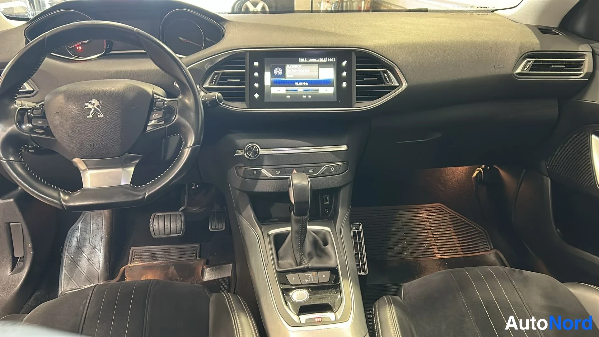 Muu: Ohjaamosta eteenpäin kuvattu näkymä: ohjauspyörä Peugeot-emblemillä, kosketusnäytöllinen infotainment-järjestelmä ja automaattivaihteiston valitsin.