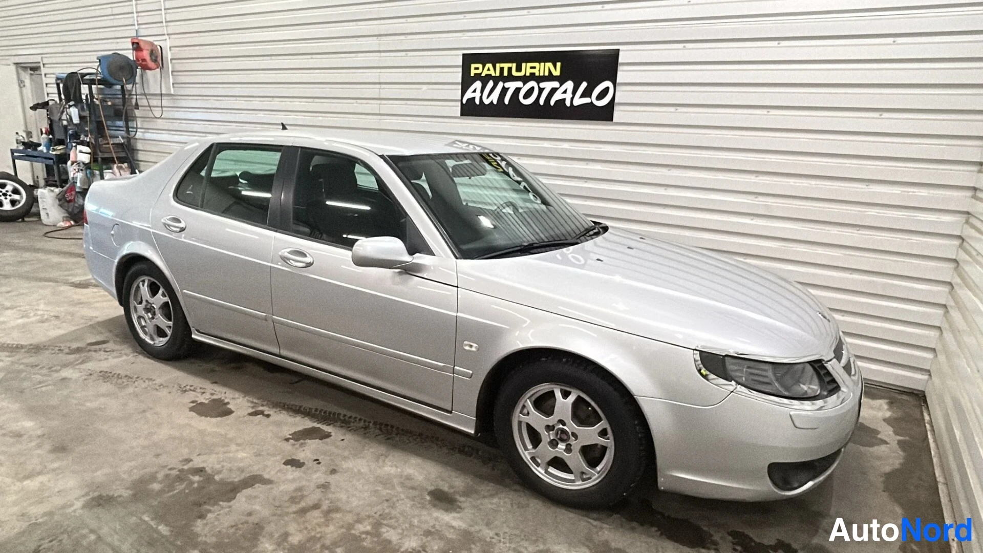Etuviisto, nokka vasemmalle: Silver Saab sedan seen from the front-right, showing the grille, headlights and driver’s side wheel. The car is inside a garage with white corrugated w