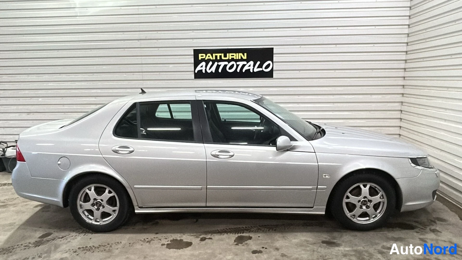 Sivulta: Full side view of a silver Saab sedan parked indoors against a white corrugated metal wall. All four doors, windows and alloy wheels are visible.