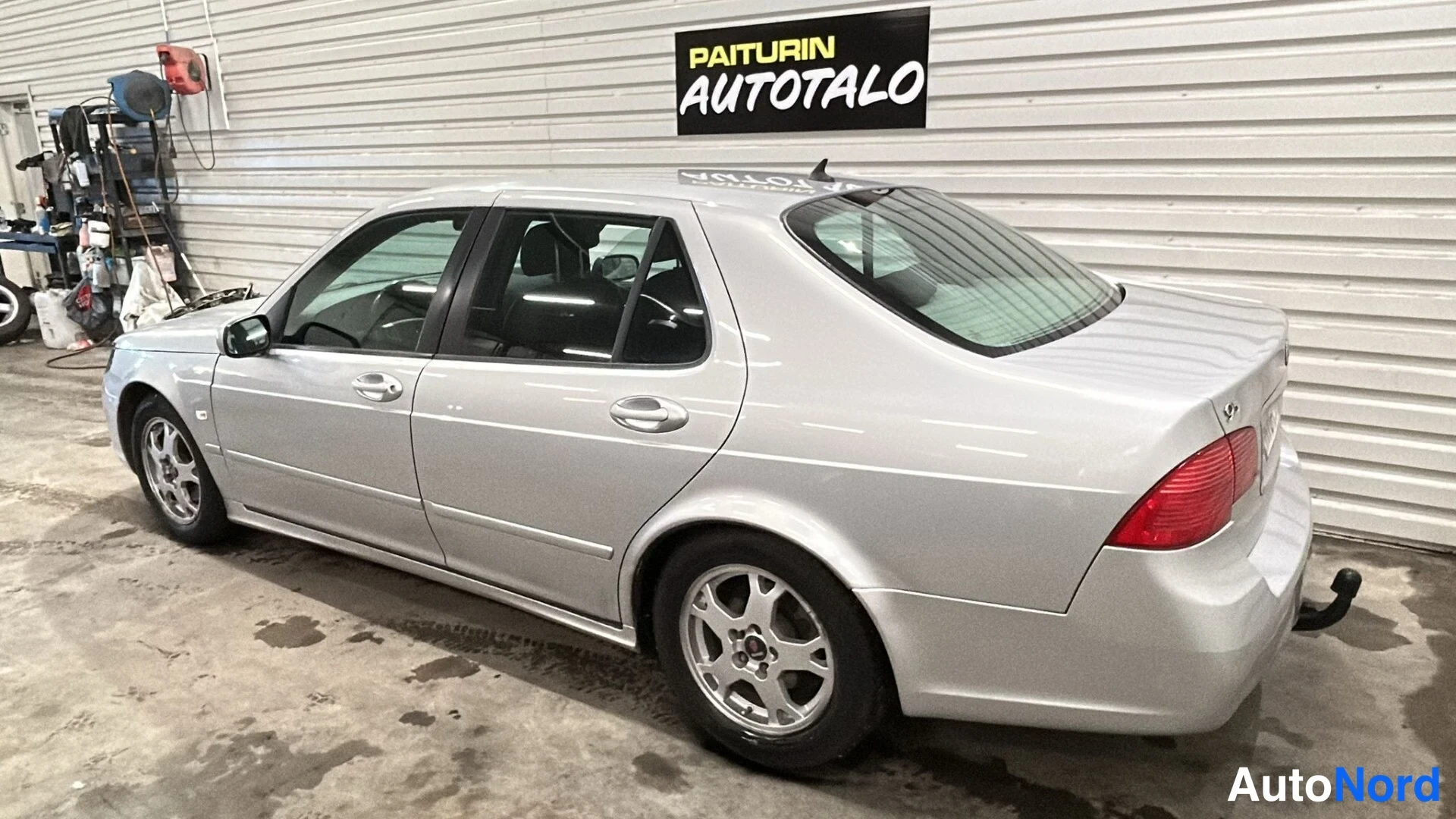 Takaviisto: Rear-left view of a silver Saab sedan in a garage, revealing the tail lamps, boot lid, left flank, rear bumper and a black tow bar fitted below the right side o