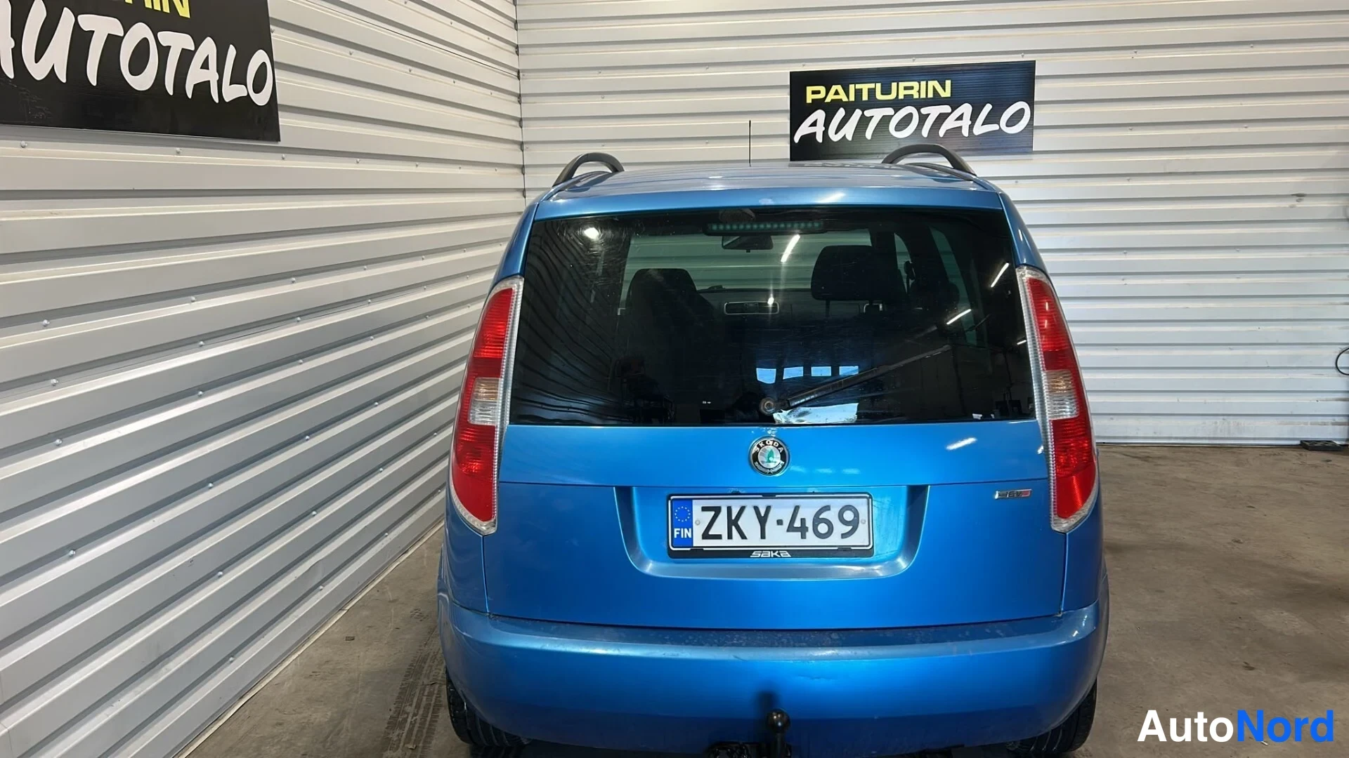 Takaa: Sininen henkilöauto suoraan takaa kuvattuna sisätilassa; takaluukku, takalasi ja takavalot näkyvät, rekisteritunnus.